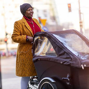 A dark woman in a cap and yellow coat stands behind a bicycle cart in an urban environment. Photo.