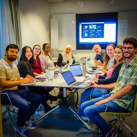 The attendees to a workshop (6 women and three men) on matlab are sitting around a table with their laptops and a screen on the background. 