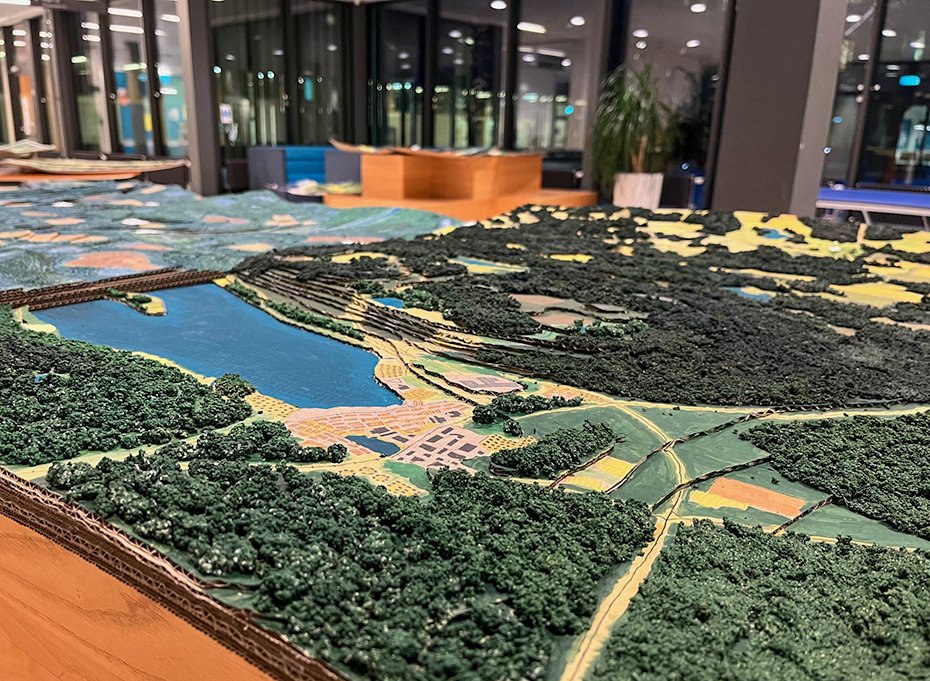 En modell gjord i wellpapp som visar landskap med vatten, bebyggelse och skog.