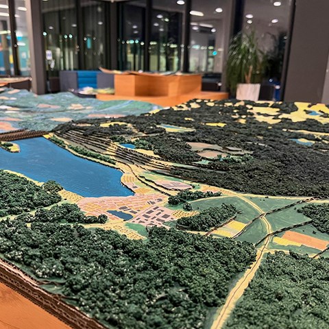 En modell gjord i wellpapp som visar landskap med vatten, bebyggelse och skog.