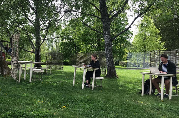 Tre skrivbord av trä på en gräsmatta. Vid två av dem sitter personer och jobbar.