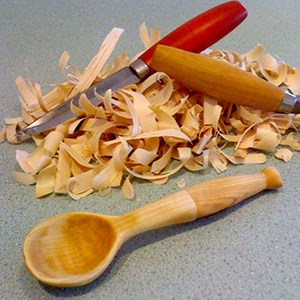 A wooden spoon, a wood carving knife, a whittling knife and wood shavings. Photo.
