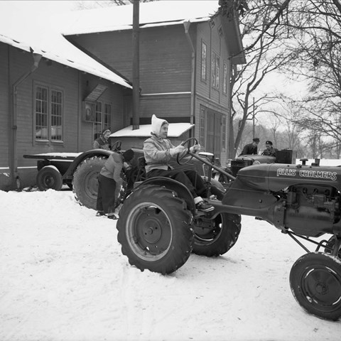 Traktor bredvid hus i snö, svartvitt fotografi. 