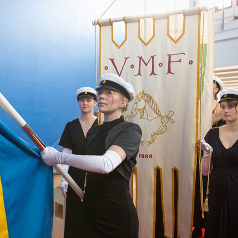 Studenter i svarta högtidskläder, vita handskar och studentmössor bär fanor i procession.