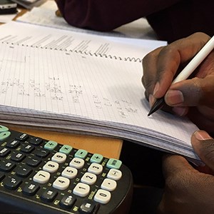 Brown hands make calculations in white notebook. Minicalculator at side. Photo.