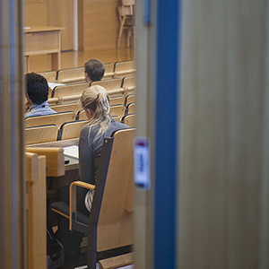 Genom halvöppen dörr skymtar studenter i universitetsaula. Foto.