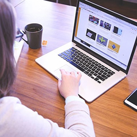 Kvinnlig student vid skrivbord använder bärbar dator, svart kaffekopp intill. Foto.