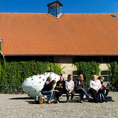 Students sit on park bench in front of house with green ivy and red tiles in Alnarp, Skåne. Photo.