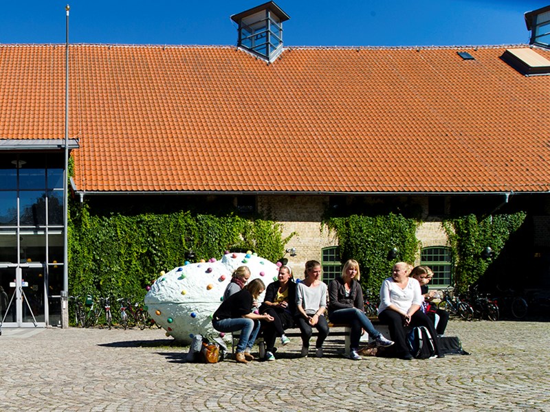 Studenter sitter på parkbänk framför stenhus med murgröna och rött tegeltak  i Alnarp, Skåne. Foto.