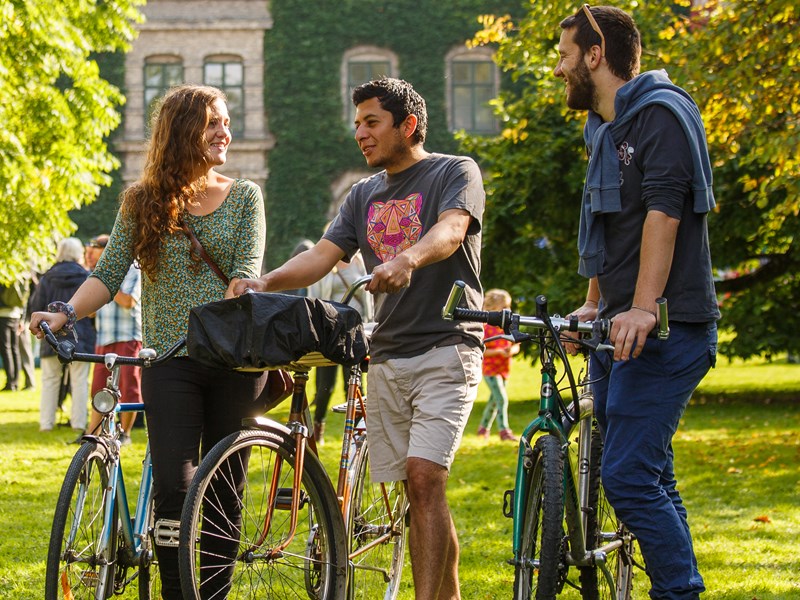 Tre studenter står med sina cyklar på campusområdet, foto.